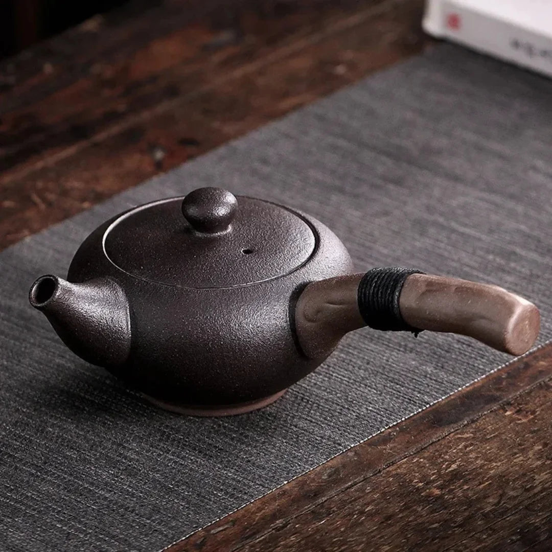 Théière japonaise en céramique noire mate avec poignée latérale en bois, posée sur un tapis.