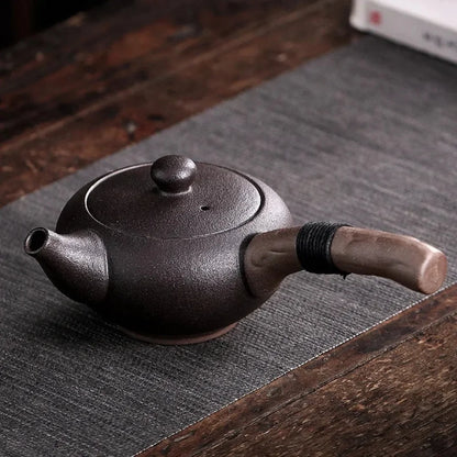 Théière japonaise en céramique noire mate avec poignée latérale en bois, posée sur un tapis.