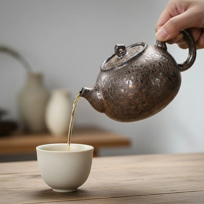Main versant le thé depuis une petite théière japonaise en céramique sombre dans une tasse blanche, sur une table en bois.