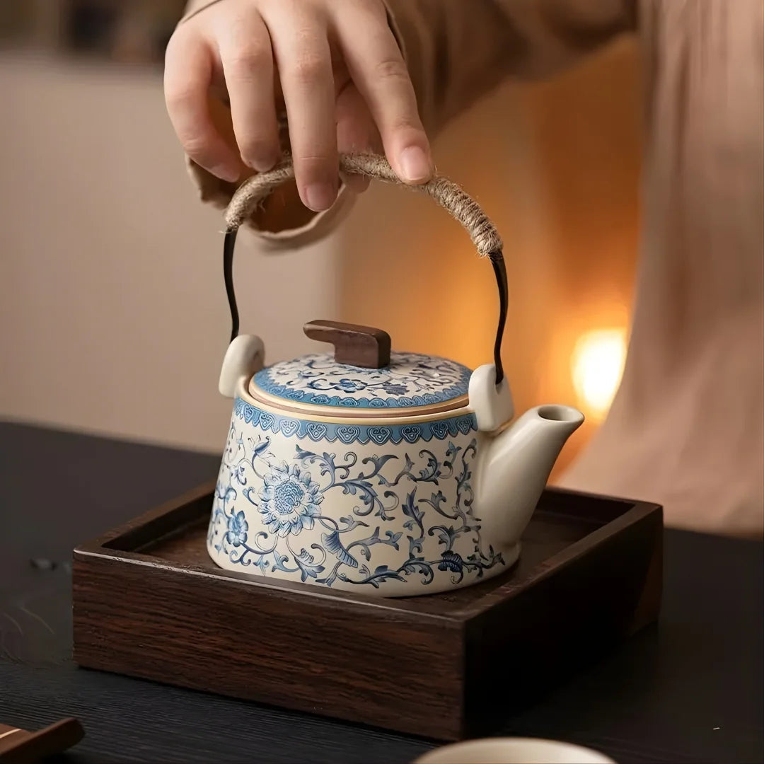 Main tenant l’anse gainée de corde d’une théière japonaise en céramique décorée, présentée sur un plateau en bois.