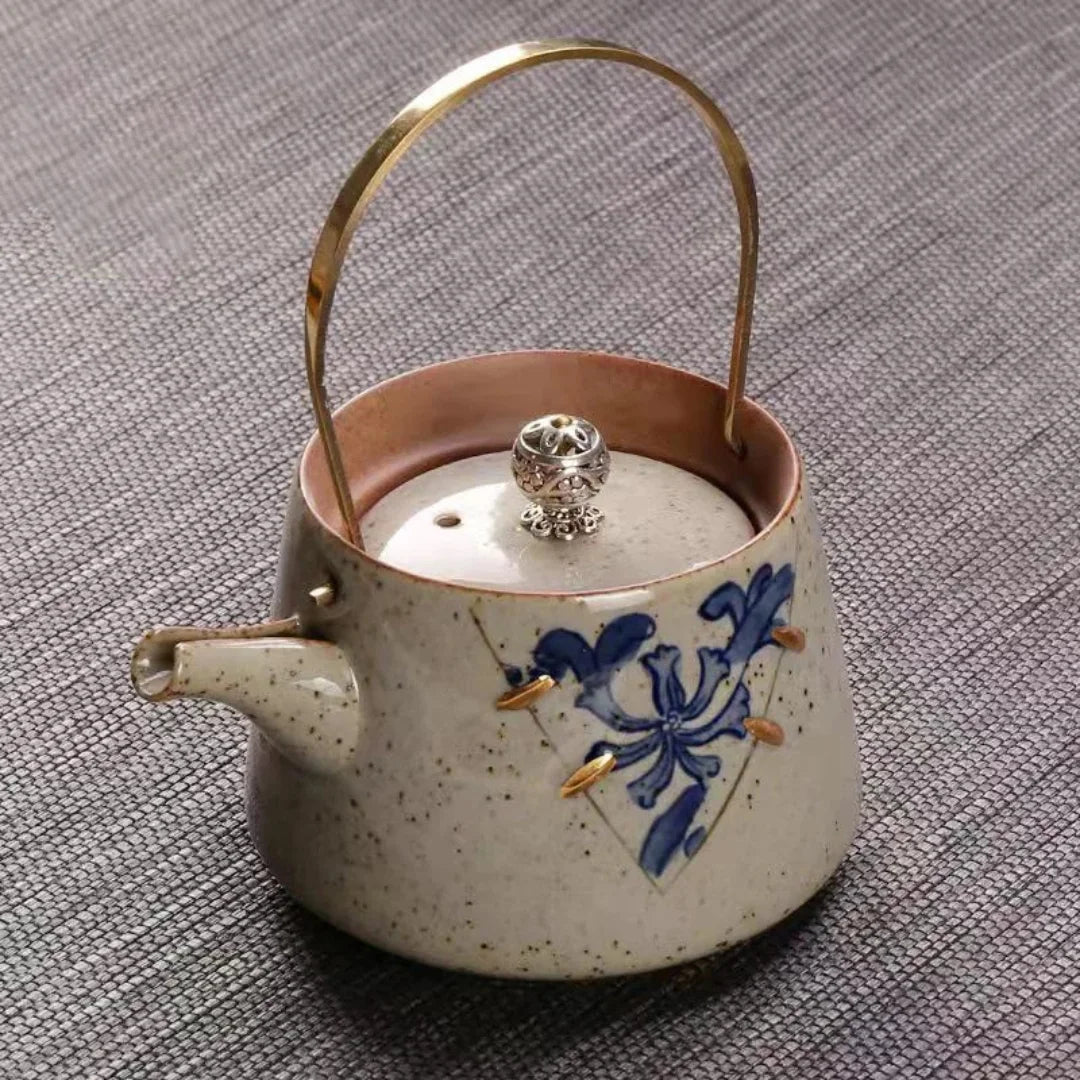 Théière artisanale en céramique crème mouchetée, décor floral bleu, anse en laiton et couvercle avec bouton sculpté.