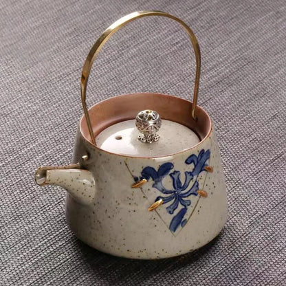 Théière artisanale en céramique crème mouchetée, décor floral bleu, anse en laiton et couvercle avec bouton sculpté.