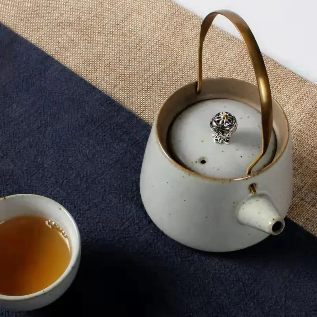 Vue de dessus d’une théière japonaise en céramique claire avec anse en laiton, posée à côté d’une tasse de thé.