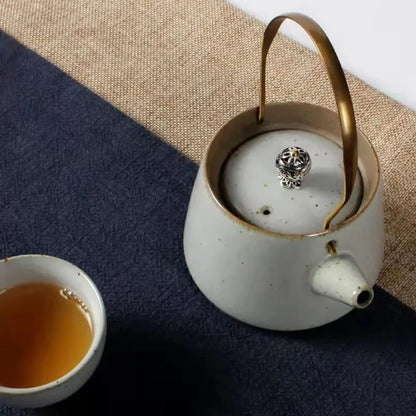 Vue de dessus d’une théière japonaise en céramique claire avec anse en laiton, posée à côté d’une tasse de thé.