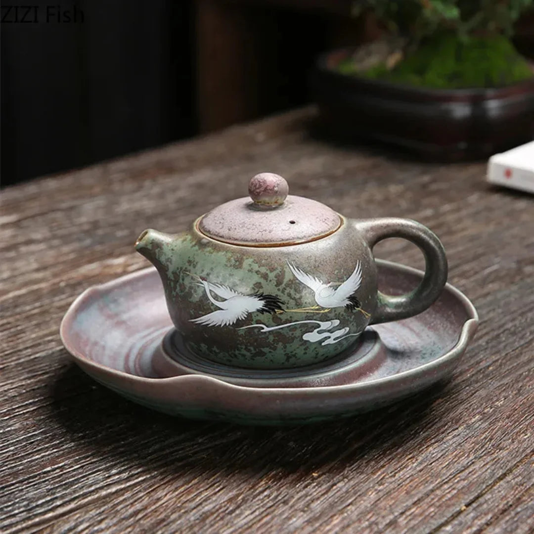 Petite théière japonaise en céramique avec motif de grues, posée sur une soucoupe assortie sur une table en bois.
