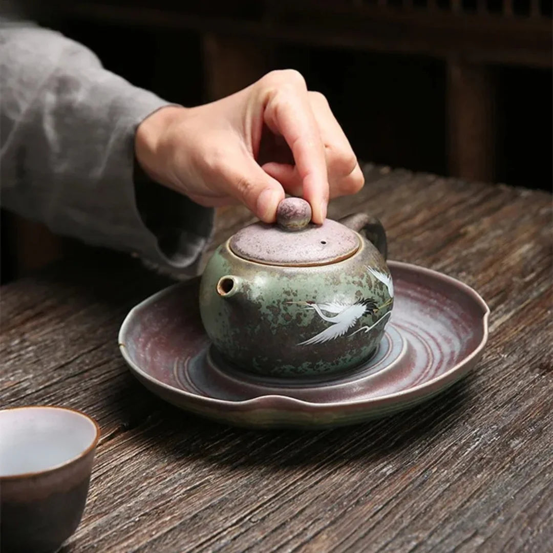 Main soulevant le couvercle d’une théière japonaise en céramique décorée de grues, placée sur une soucoupe.