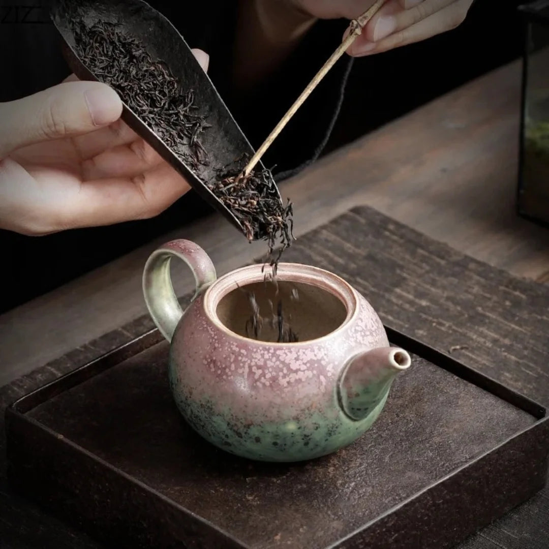 Des feuilles de thé sont versées dans une petite théière japonaise en céramique émaillée aux nuances vert et rose, posée sur un plateau.