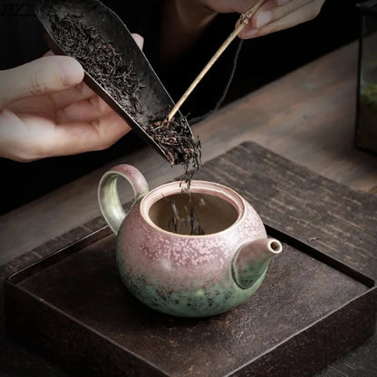 Des feuilles de thé sont versées dans une petite théière japonaise en céramique émaillée aux nuances vert et rose, posée sur un plateau.