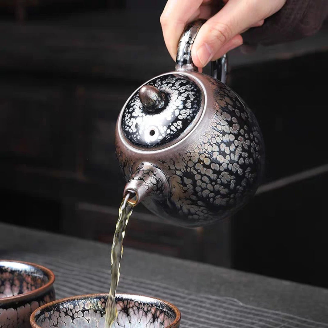 Main versant du thé depuis une théière japonaise à glaçure cristallisée noire et argentée dans des tasses assorties.